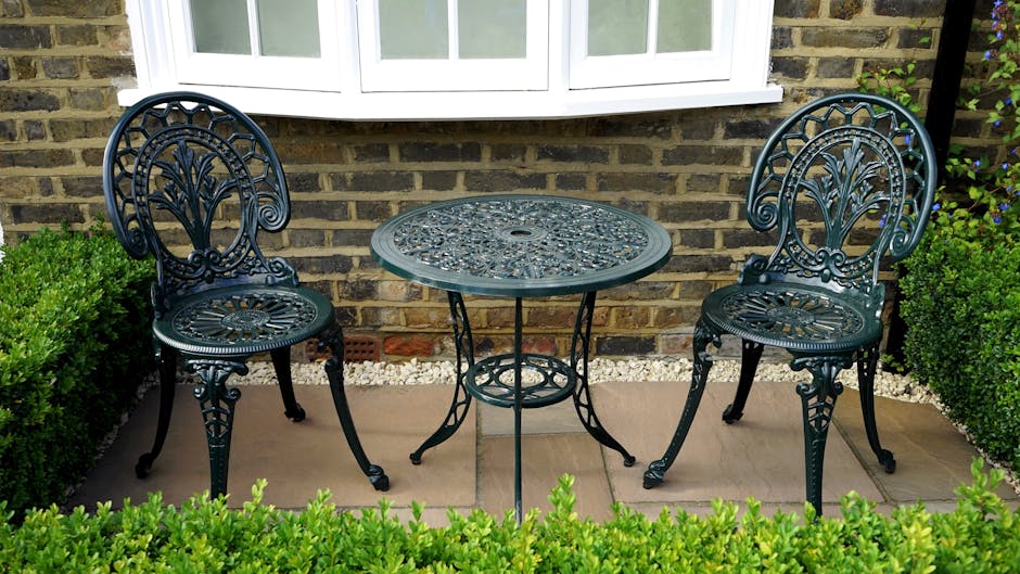 A small front garden area featuring a decorative wrought iron table with an intricate circular pattern, flanked by two matching wrought iron chairs with ornate backrests and curved legs. The garden is