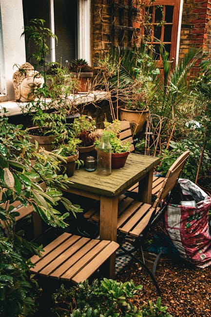A cozy urban garden corner with a weathered wooden outdoor table surrounded by matching wooden benches and chairs, set against a brick wall with a window. The table is adorned with small potted plants